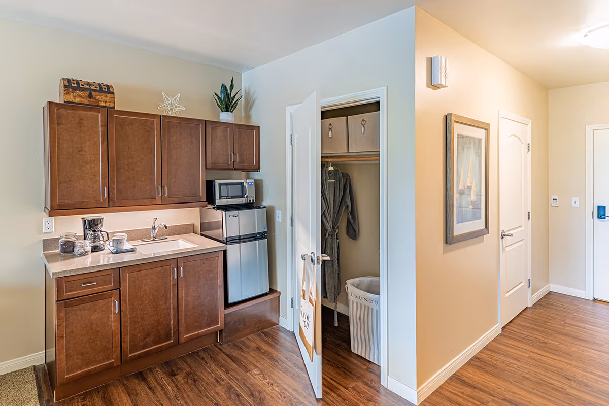 Small kitchenette with wooden cabinets, a sink, microwave and mini fridge next to an open closet and hallway.