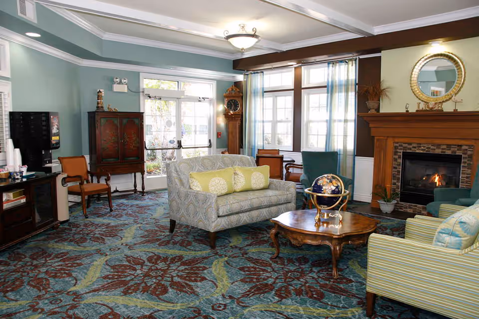 A cozy living room area with a patterned carpet, a fireplace with a wooden mantel, a round wooden coffee table with a decorative globe, a loveseat with green and white pillows, several armchairs, a grandfather clock, and large windows with sheer curtains letting in natural light.