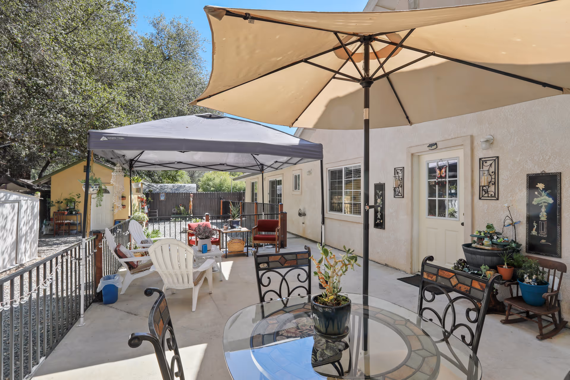 Outdoor patio area at Citrus Pines Senior Living with a glass table and metal chairs under a large beige umbrella. There are additional seating areas with white plastic chairs and red cushioned chairs under a gray canopy. The patio is surrounded by a black metal fence, and there are plants and decorative wall hangings on the building's exterior wall. Trees and a small yellow shed are visible in the background.