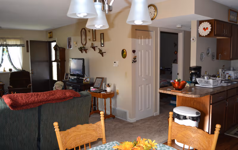 Interior view of a senior living facility showing a combined living room and kitchen area. The living room has a green couch with a red blanket draped over it, a TV on a stand, and wall decorations including clocks and small shelves. The kitchen area features wooden cabinets, a countertop with a fruit bowl, a coffee maker, and a dish drying rack. A wooden dining table with chairs and a floral centerpiece is partially visible in the foreground. There is a doorway leading to another room and a window with curtains letting in natural light.