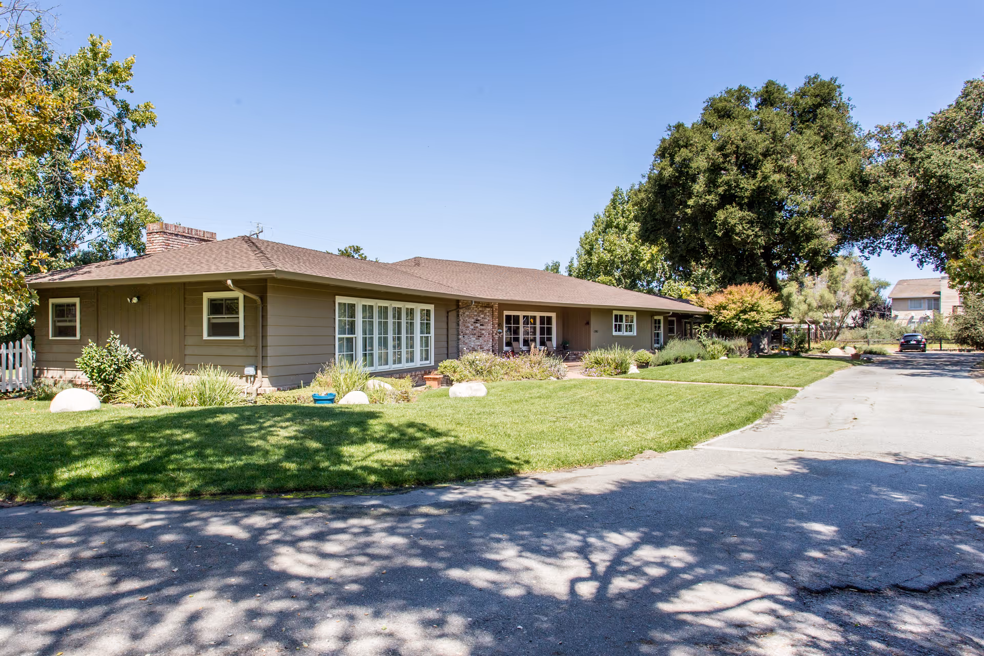 Single-story ranch-style senior care building with brown siding, a manicured front lawn, driveway, and large trees under a clear blue sky.