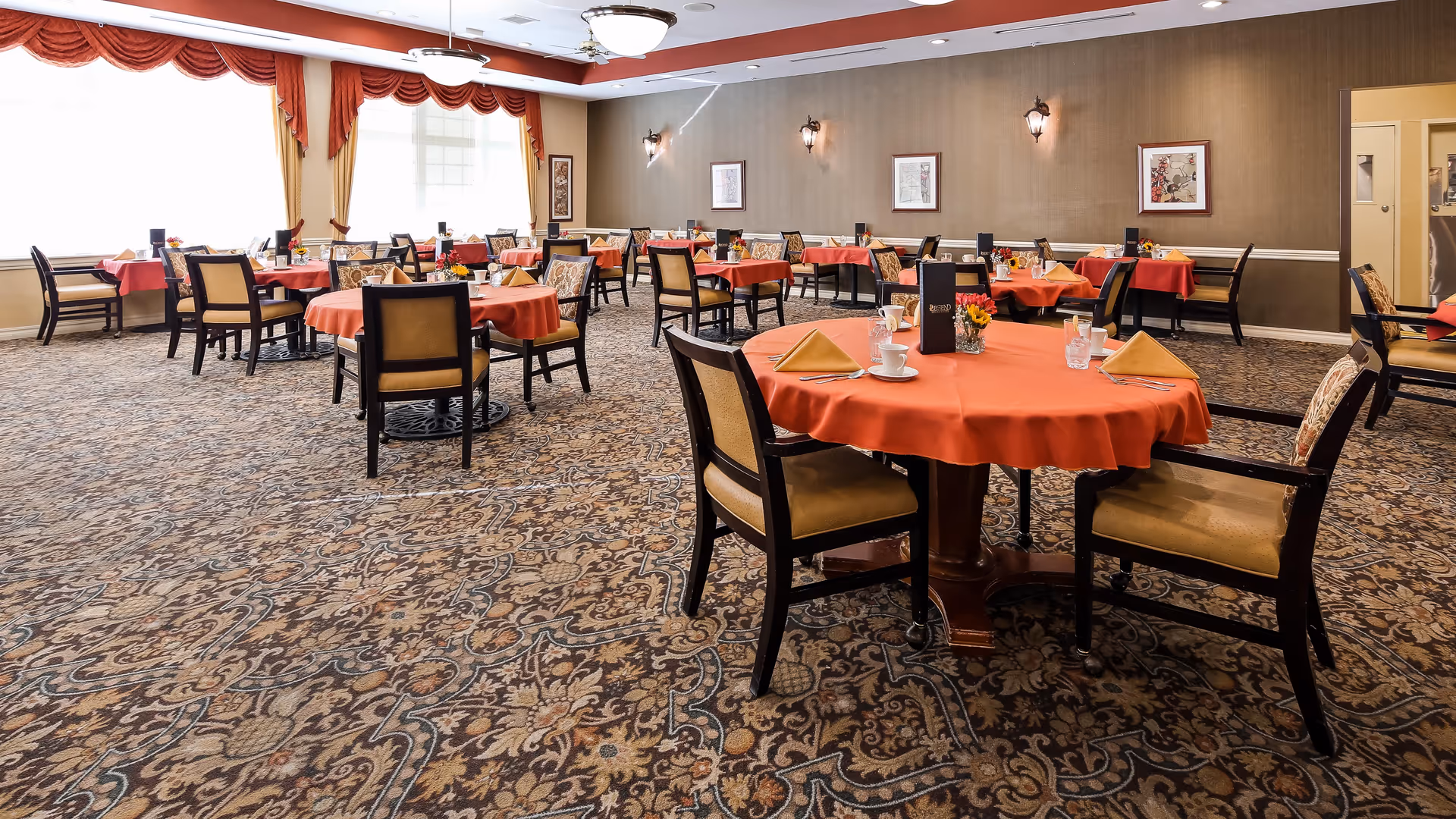 A spacious dining room with multiple round tables covered in orange tablecloths, each set with napkins, cups, and small flower arrangements. The room features patterned carpet, beige walls with framed artwork, large windows with red and gold curtains, and ceiling light fixtures.