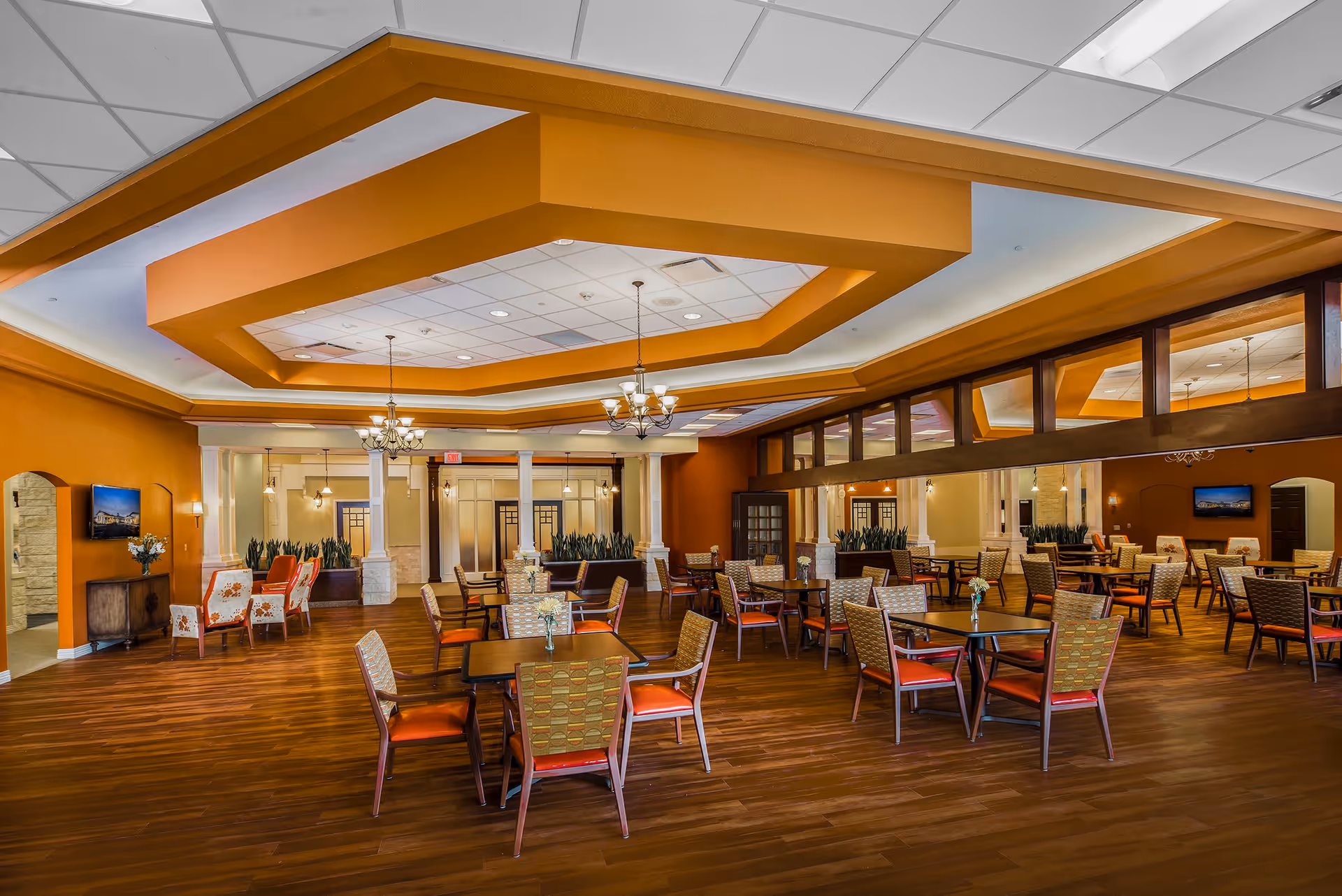 Spacious dining room with multiple tables and chairs arranged neatly on a wooden floor. The ceiling features a large, decorative recessed design with chandeliers hanging down. The walls are painted in warm tones of orange and beige, with large mirrors and windows adding depth to the room. There are some plants and a TV mounted on the wall, creating a welcoming atmosphere.