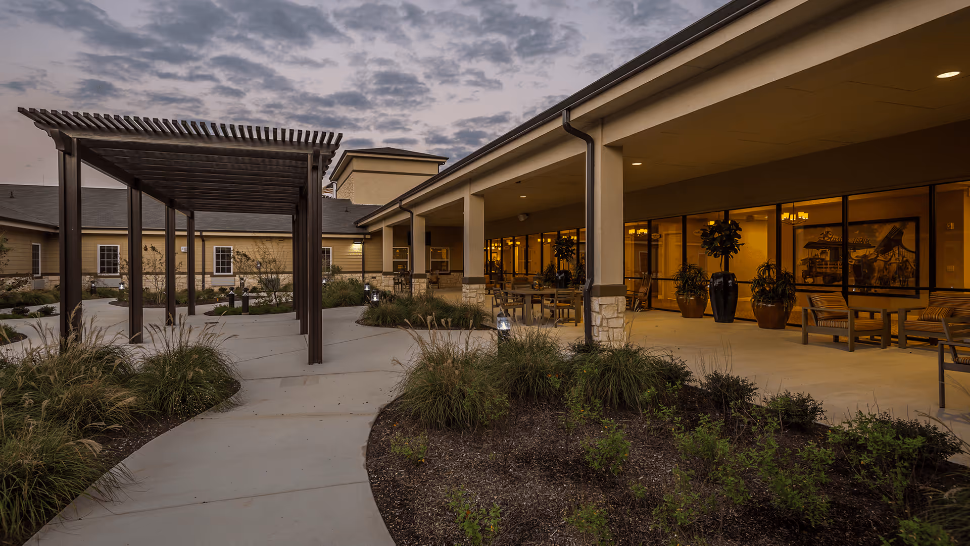 Outdoor patio area at dusk with a covered walkway, landscaped garden beds, and seating arrangements outside a building with large windows showing indoor lighting.