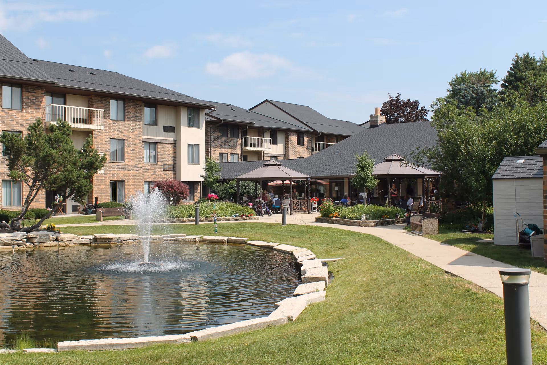Outdoor view of a senior living facility with a pond and fountain in the foreground, surrounded by a stone border and green grass. In the background, there are multi-story brick and beige buildings with balconies, and a covered patio area with people sitting and socializing. Trees and shrubs are also visible around the area.