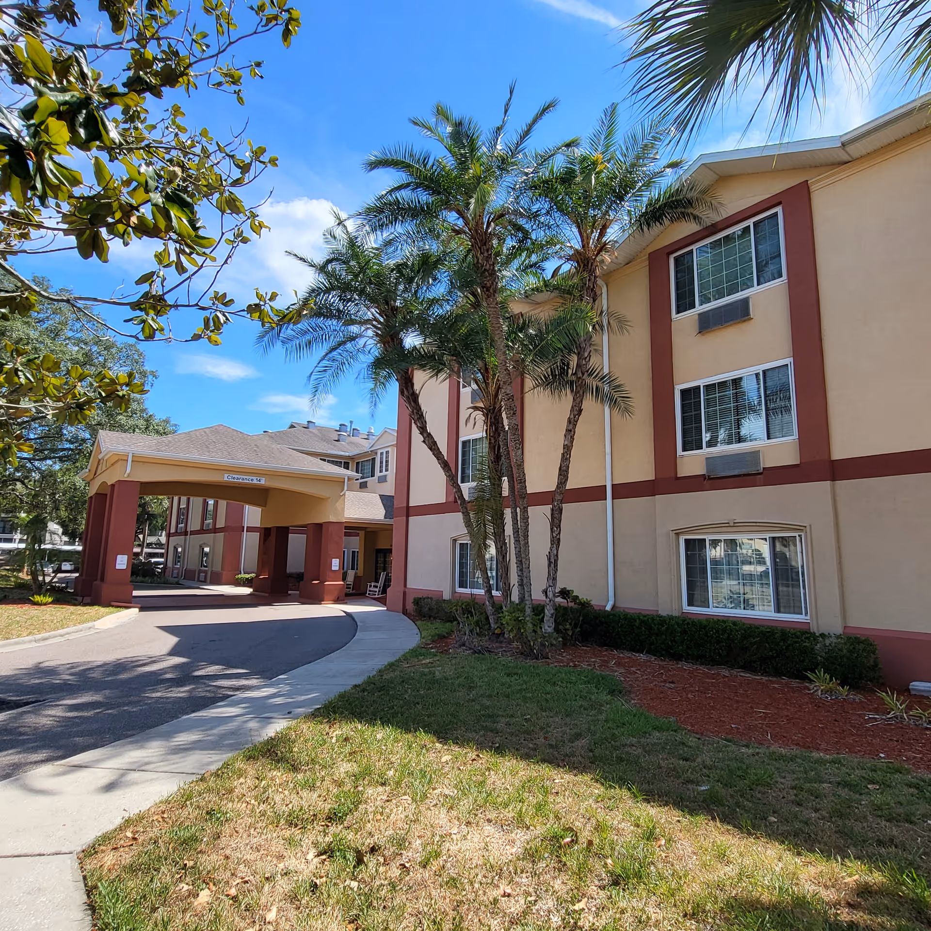 Front exterior of a three-story beige-and-red senior living building with palm trees, an entrance canopy and a curved driveway.