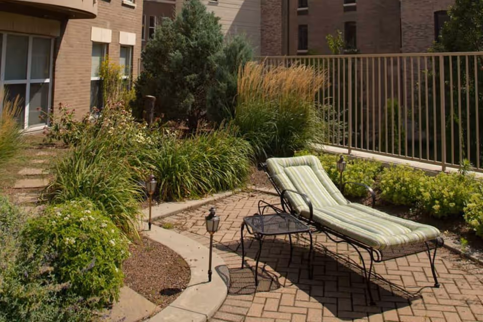 Outdoor patio area with a cushioned lounge chair and a small metal table surrounded by various green plants and bushes, with a building and railing in the background.