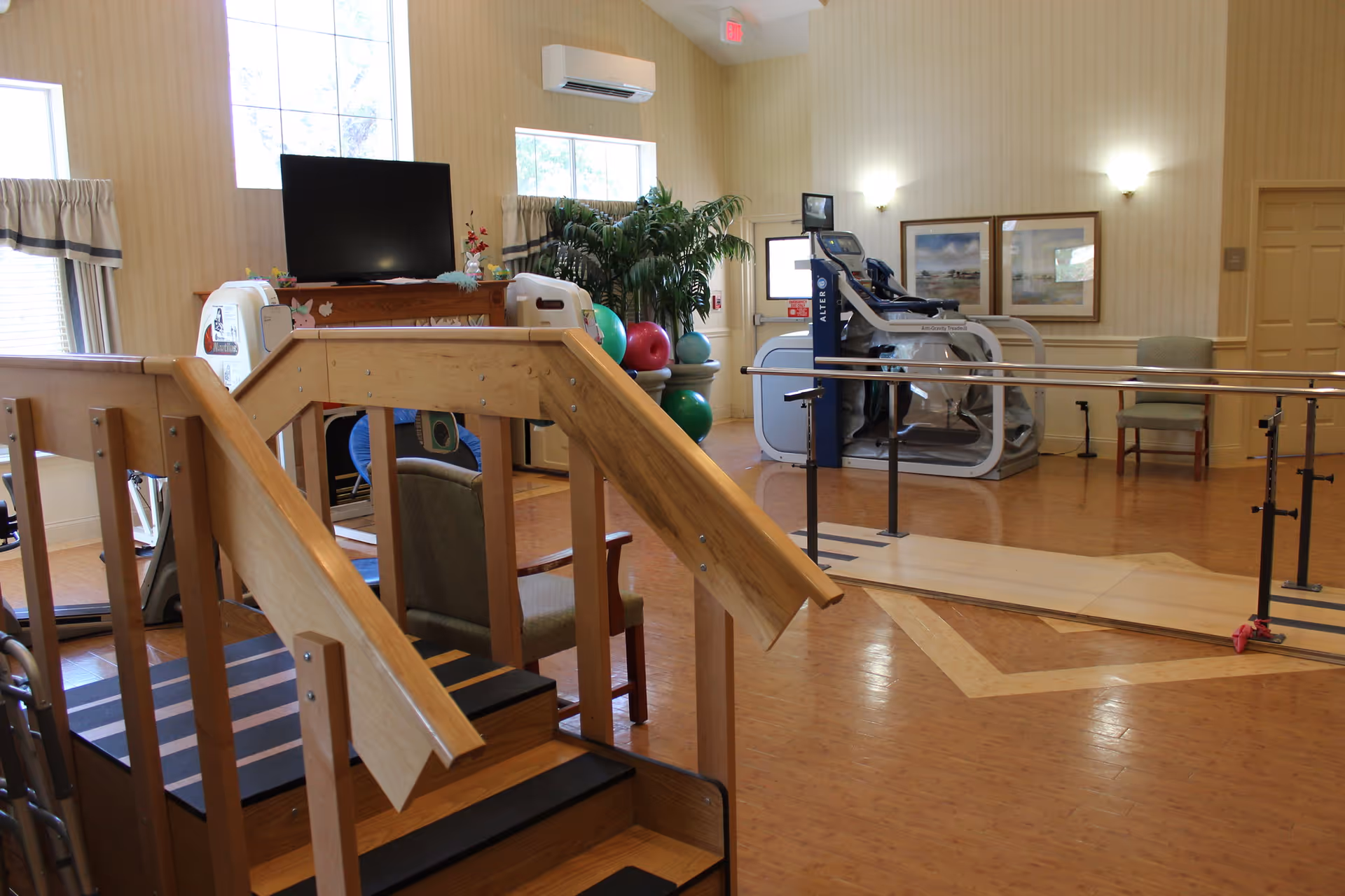 Interior view of a rehabilitation center room featuring wooden stairs with handrails for physical therapy, parallel bars for walking exercises, exercise equipment including a treadmill, a television mounted on a wooden stand, chairs, and large windows with curtains allowing natural light.