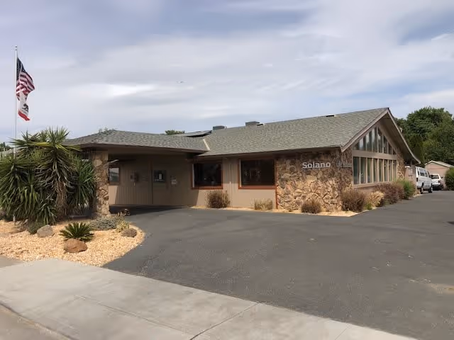 Exterior view of a single-story building with stone and beige siding, a sloped roof, and large windows. There are two flagpoles with the American and California flags near the entrance. The building is surrounded by a paved driveway and some landscaping with rocks and plants.