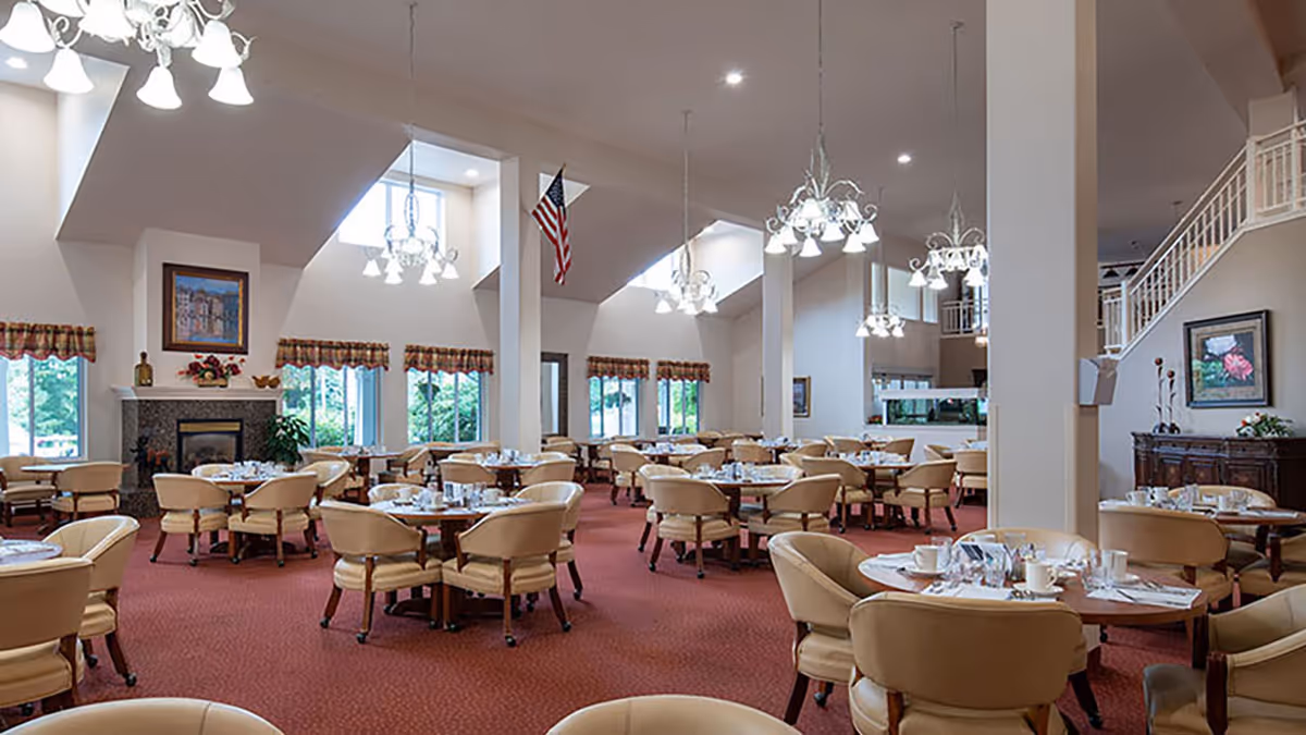 Large dining room with round tables set for meals, beige chairs, chandeliers, and high vaulted ceilings.