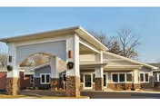 Exterior view of a single-story healthcare facility building with a covered entrance supported by stone pillars. The building has a light-colored facade with multiple windows and a clear sky in the background.