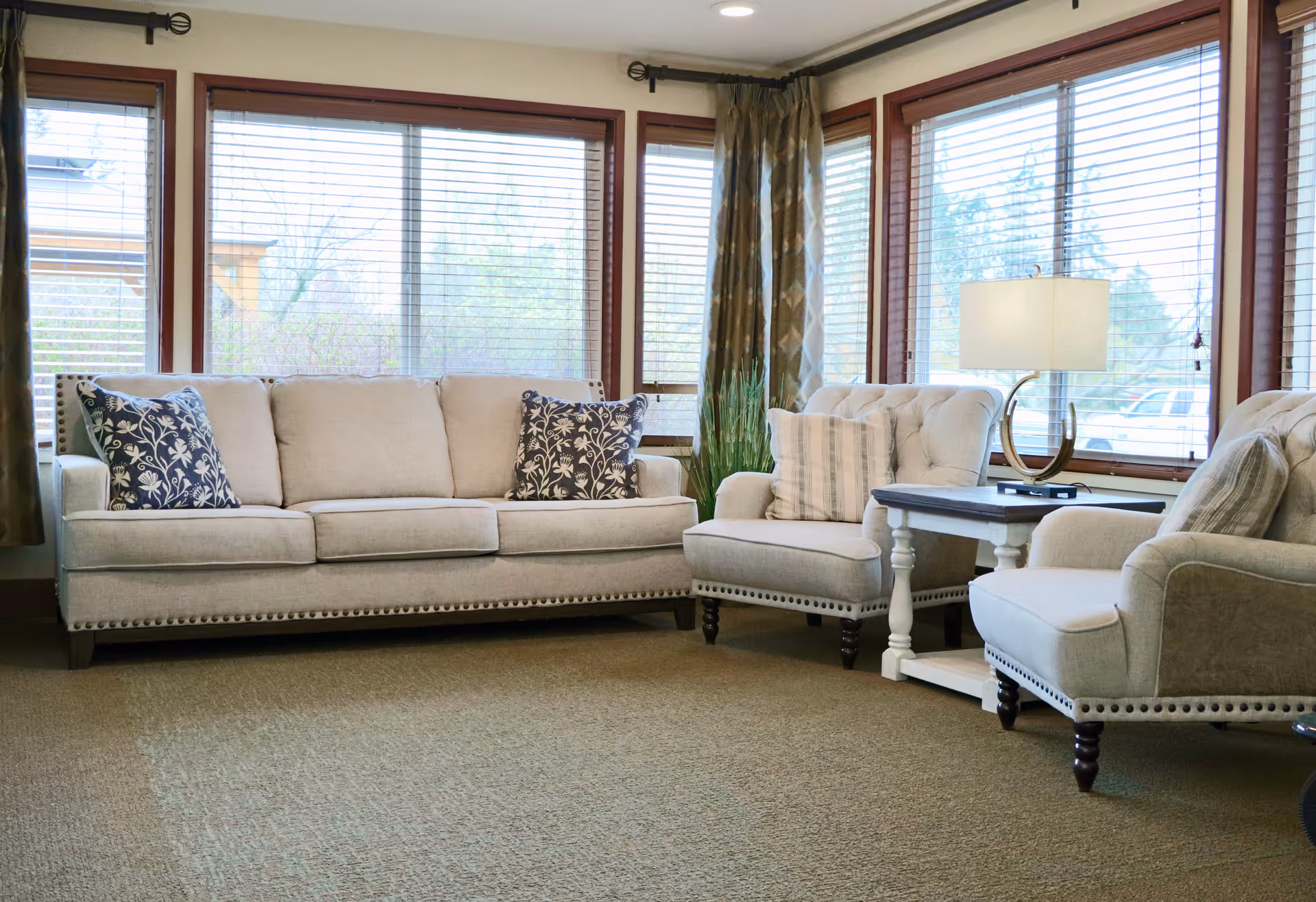 A cozy living room with a beige sofa and two matching armchairs, each adorned with decorative pillows. A small white side table with a modern lamp sits between the armchairs. Large windows with wooden blinds and patterned curtains allow natural light to fill the room.