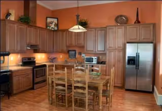 A spacious kitchen with wooden cabinets, a stainless steel refrigerator, an oven, microwave, and a wooden dining table with six chairs in the center. The walls are painted a warm orange color, and there are decorative items on top of the cabinets.