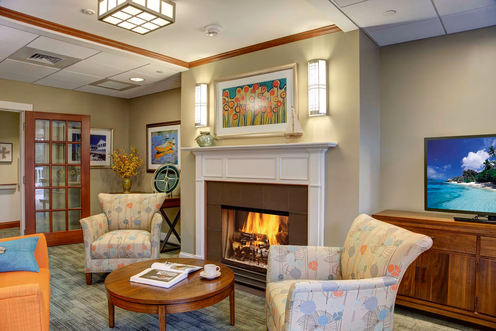 A cozy living room area in a senior living facility featuring a lit fireplace with a white mantel, two patterned armchairs, an orange sofa with a blue pillow, a round wooden coffee table with a book and a cup, a wooden cabinet with a TV displaying a beach scene, and decorative wall art and lighting.