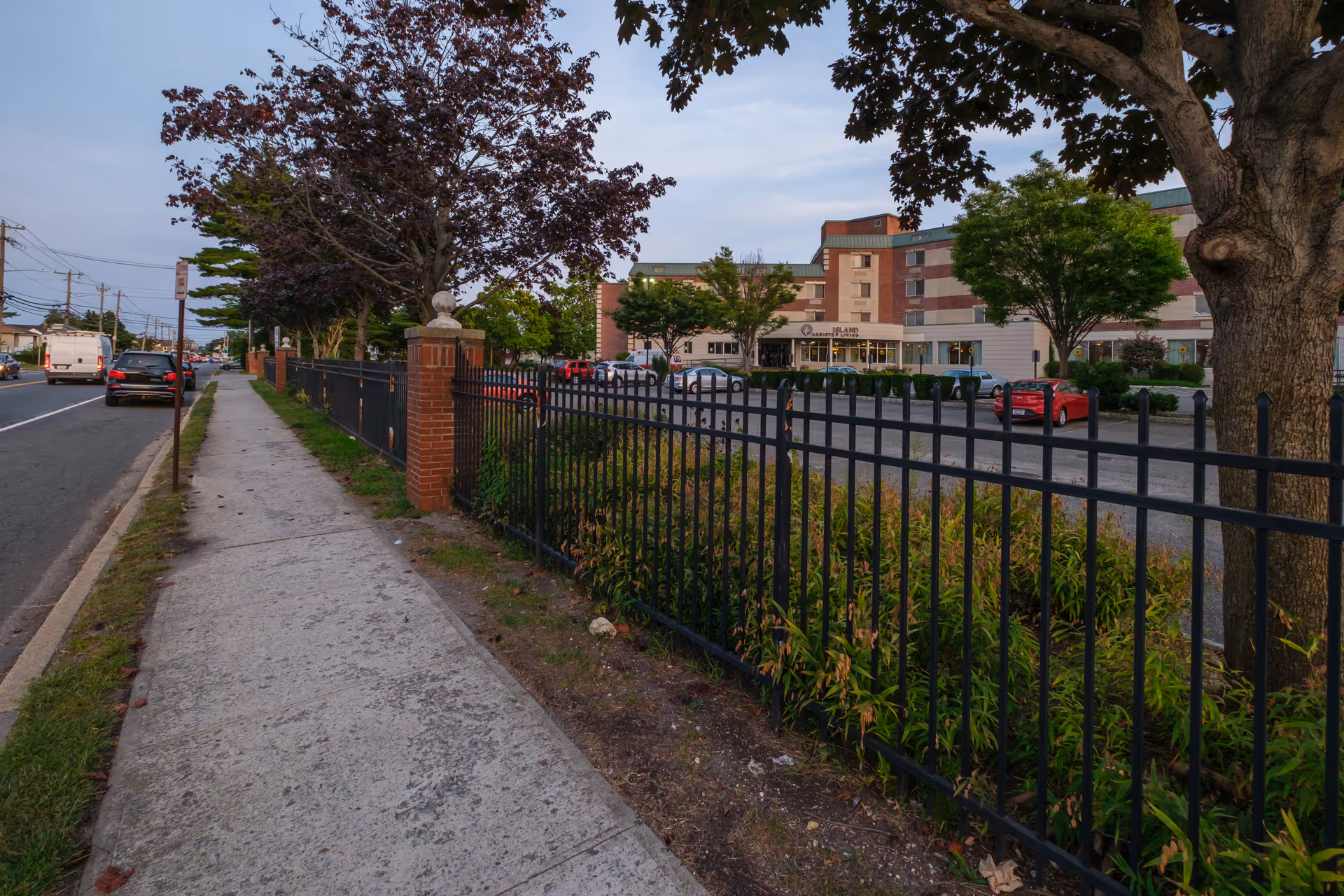Sidewalk along a street with cars parked and driving, bordered by a black metal fence with brick pillars and trees. In the background, a multi-story building with a sign reading 'Island Assisted Living' is visible, along with a parking lot containing several cars.