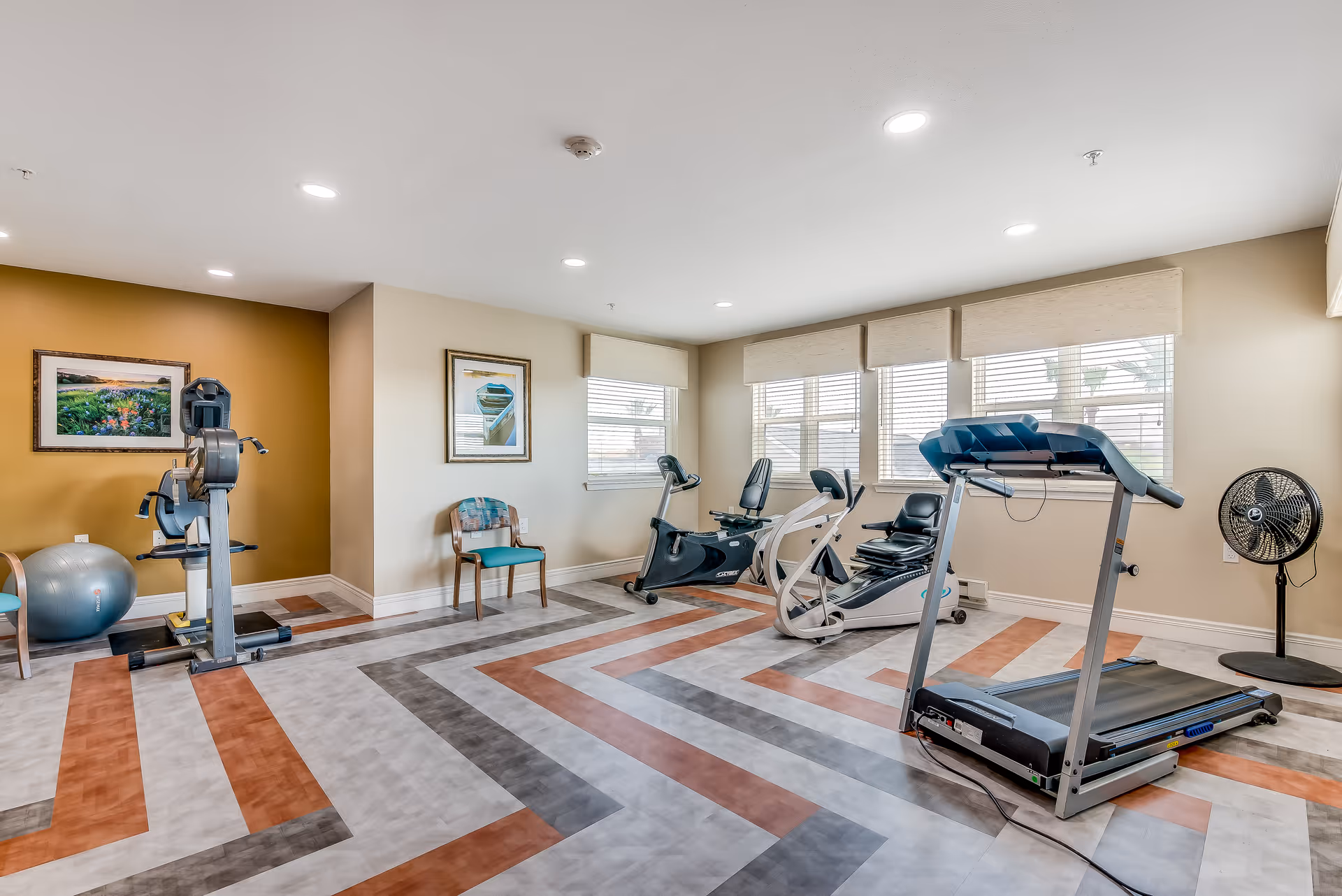 A bright exercise room with various fitness equipment including a treadmill, stationary bikes, and an exercise ball. The room has large windows with blinds, two framed pictures on the walls, and a patterned floor with orange, gray, and white colors. There are also two chairs placed against the walls.