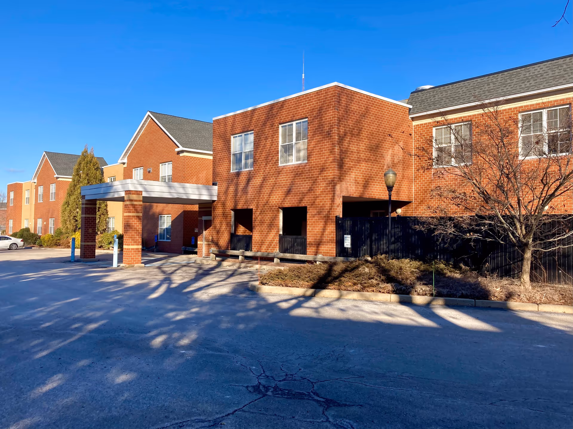 Front entrance of a red-brick senior living building with a covered porte-cochère, driveway, and leafless trees under a clear blue sky.