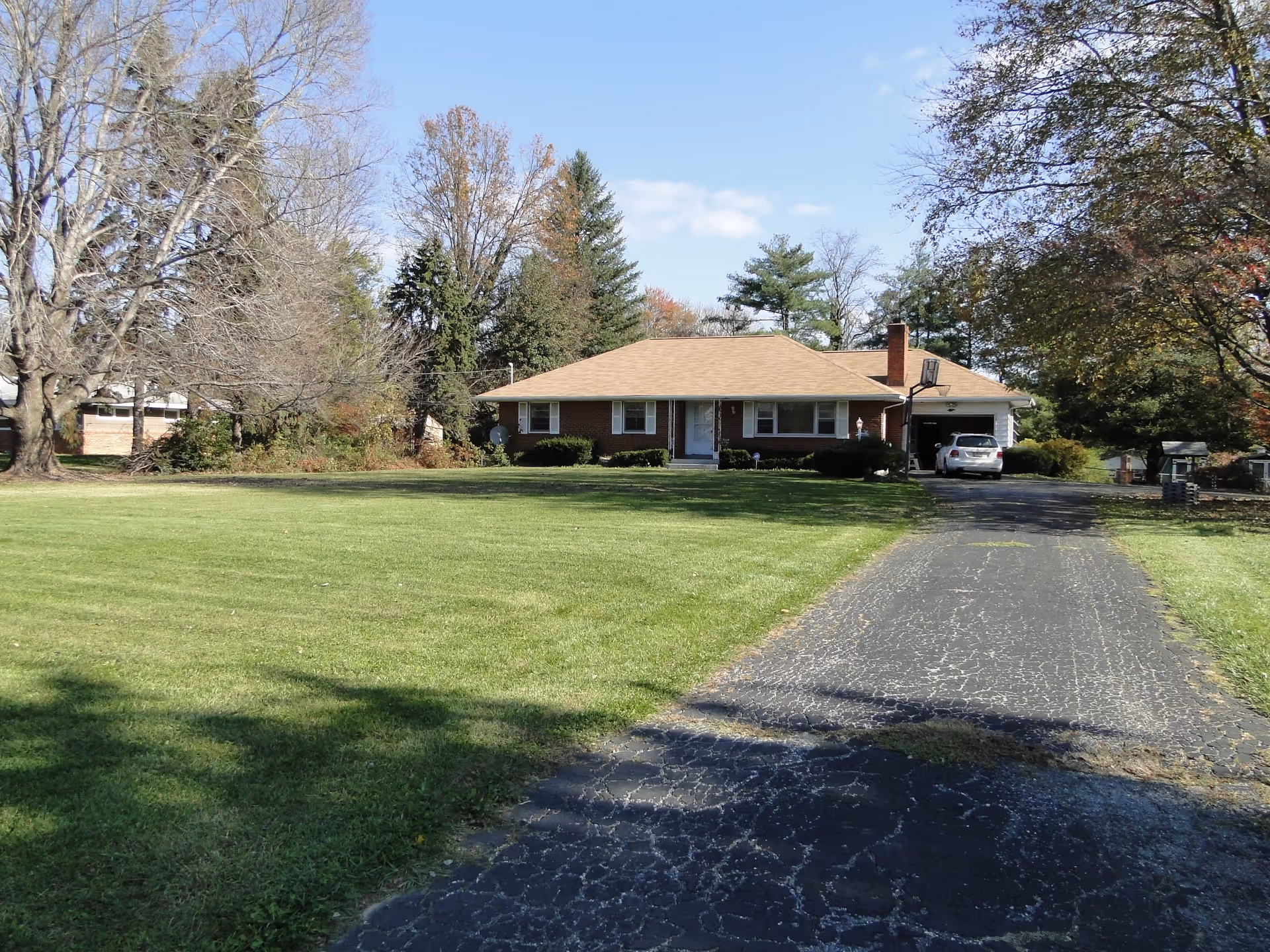 A single-story brick house with a tan roof, surrounded by a large green lawn and trees with some autumn foliage. A paved driveway leads up to the house where a white car is parked near the garage. The sky is clear with a few clouds.