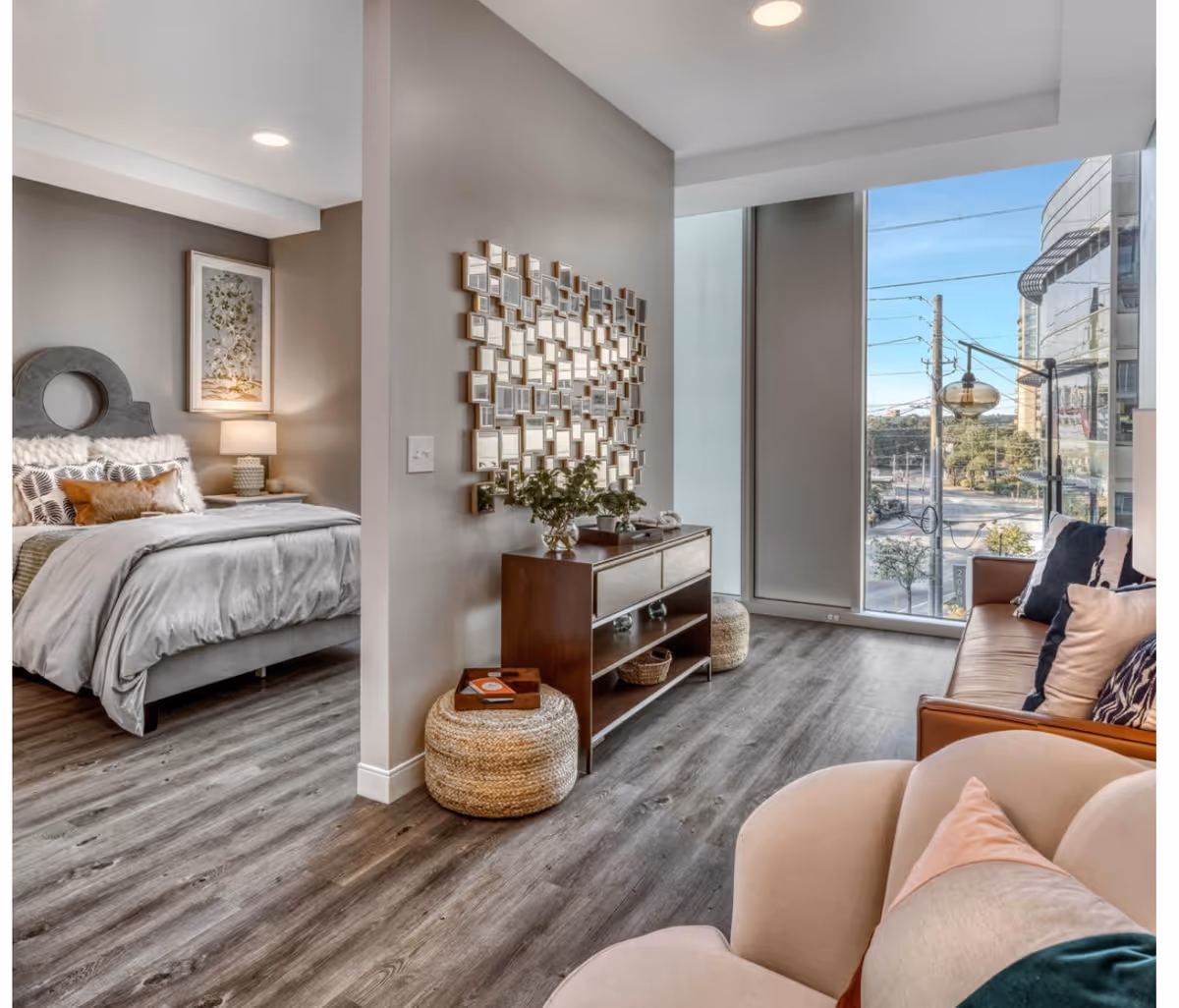 A modern senior living apartment interior featuring a cozy bedroom with a gray upholstered bed, decorative pillows, and a bedside lamp. Adjacent to the bedroom is a living area with a brown leather sofa, a beige armchair with colorful cushions, a wooden console table with plants and decor, and a large window offering a view of the city outside.