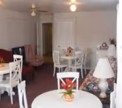 Interior view of a senior living facility common area with several white round tables and chairs, a floral patterned sofa with a white lamp on a side table, a pink armchair, and a small dining table with a flower centerpiece. The walls are light-colored with a door and hallway visible in the background.