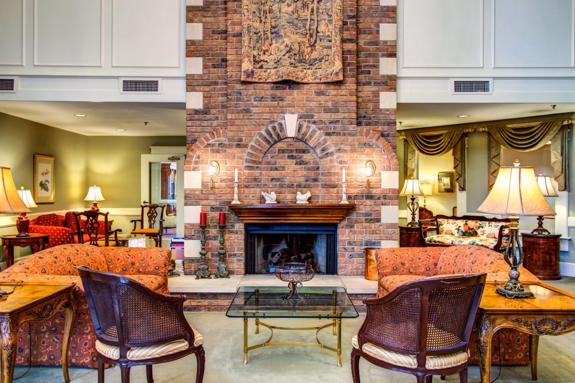 A cozy living room area with a large brick fireplace in the center. The room features two orange patterned sofas facing each other with a glass coffee table in between. There are two wooden chairs with cane backs and striped cushions in front of the sofas. The room is warmly lit with several table lamps and wall sconces. Decorative items, including candles and small sculptures, are placed on the fireplace mantel. The walls are painted light green with white paneling, and there are framed artworks and draped curtains in the background.