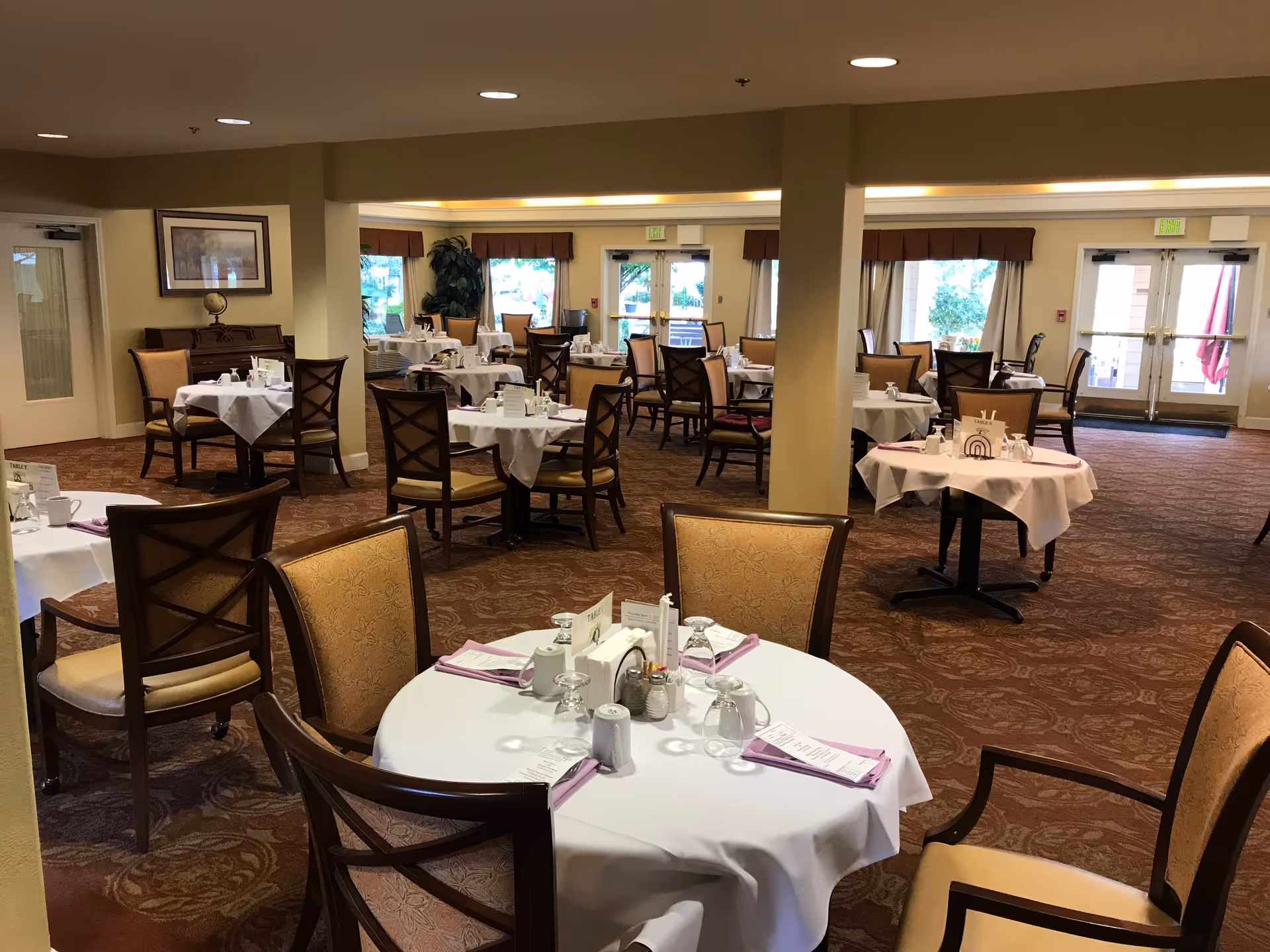 A spacious dining room with multiple round tables covered with white tablecloths, each set with napkins, glasses, and condiments. The room has beige walls, carpeted floors, large windows with curtains, and several wooden chairs with cushioned seats around the tables.