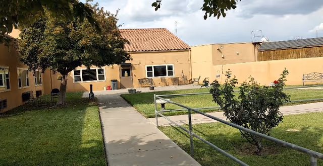 Outdoor courtyard with a concrete pathway, grassy lawn, benches, a tree, and a single-story building with a tiled roof.