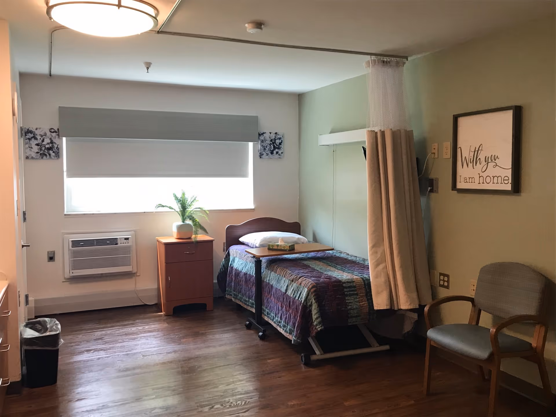 A small, cozy bedroom in a senior living facility with a single bed covered by a colorful quilt. Next to the bed is a wooden nightstand with a potted plant on top. A window with a closed blind is above the nightstand. There is a beige privacy curtain partially drawn around the bed. A framed sign on the wall reads 'With you I am home.' A wooden chair with armrests is positioned near the bed. The floor is wooden, and a ceiling light fixture is visible.