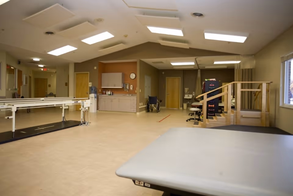 A spacious rehabilitation therapy room with parallel bars, treatment tables, a wheelchair, and exercise stairs in a senior facility.