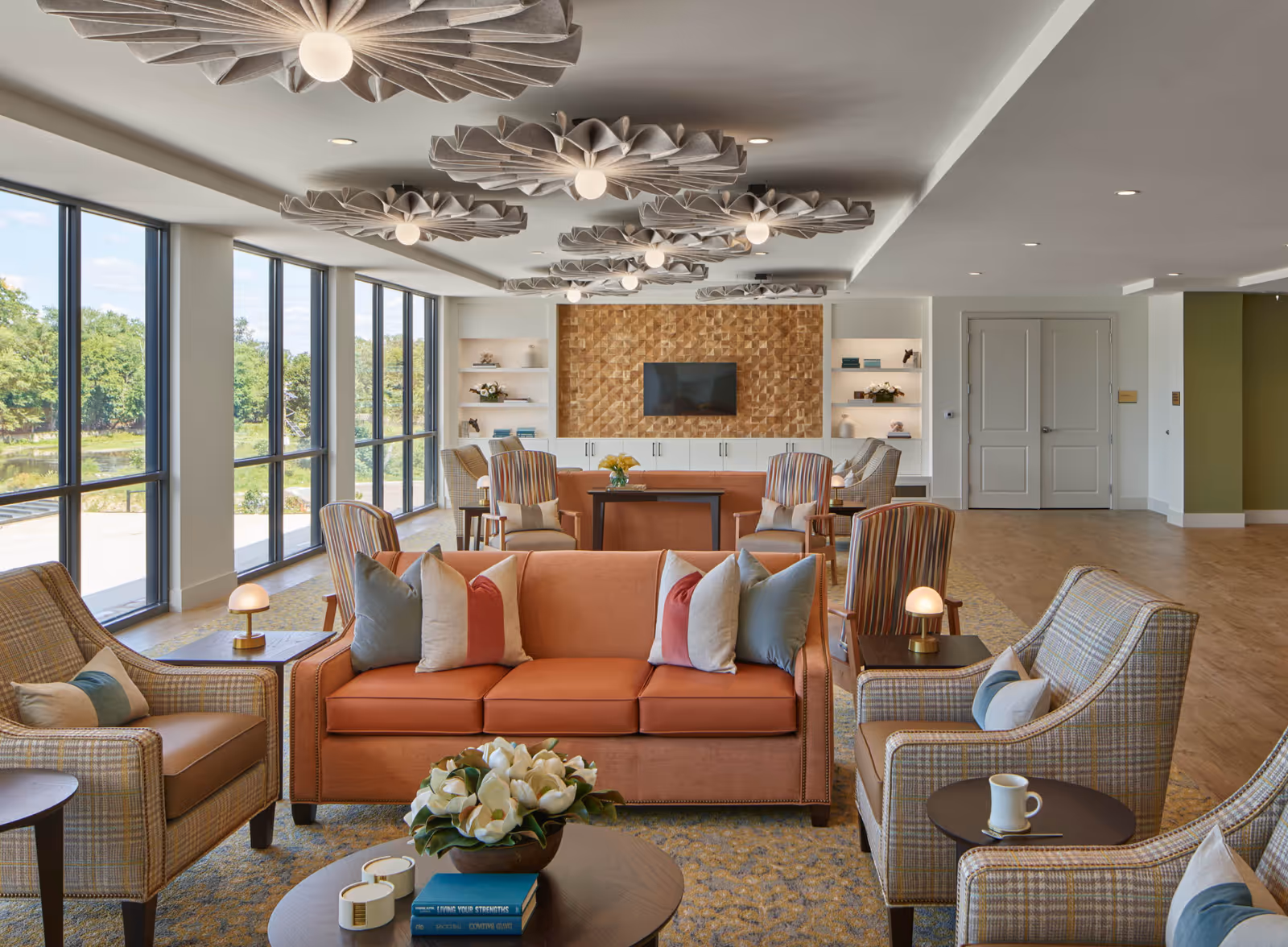 A spacious and well-lit living room area with large floor-to-ceiling windows on the left side showing greenery outside. The room features multiple seating arrangements including an orange sofa with several pillows, plaid armchairs, and small tables with lamps and a coffee cup. The ceiling has unique decorative light fixtures, and a wooden textured wall with a mounted flat-screen TV is visible at the far end of the room.