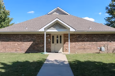Front view of a single-story brick building with a centered doorway under a small gabled porch and a concrete walkway across a grassy lawn.