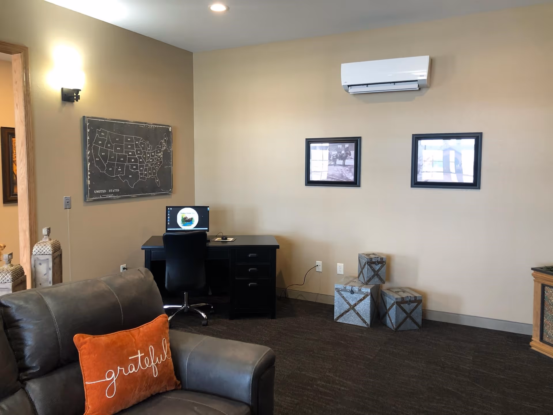 A cozy interior lounge with a leather sofa and orange 'grateful' pillow, a desk with a computer, wall art, and decorative boxes against a beige wall.