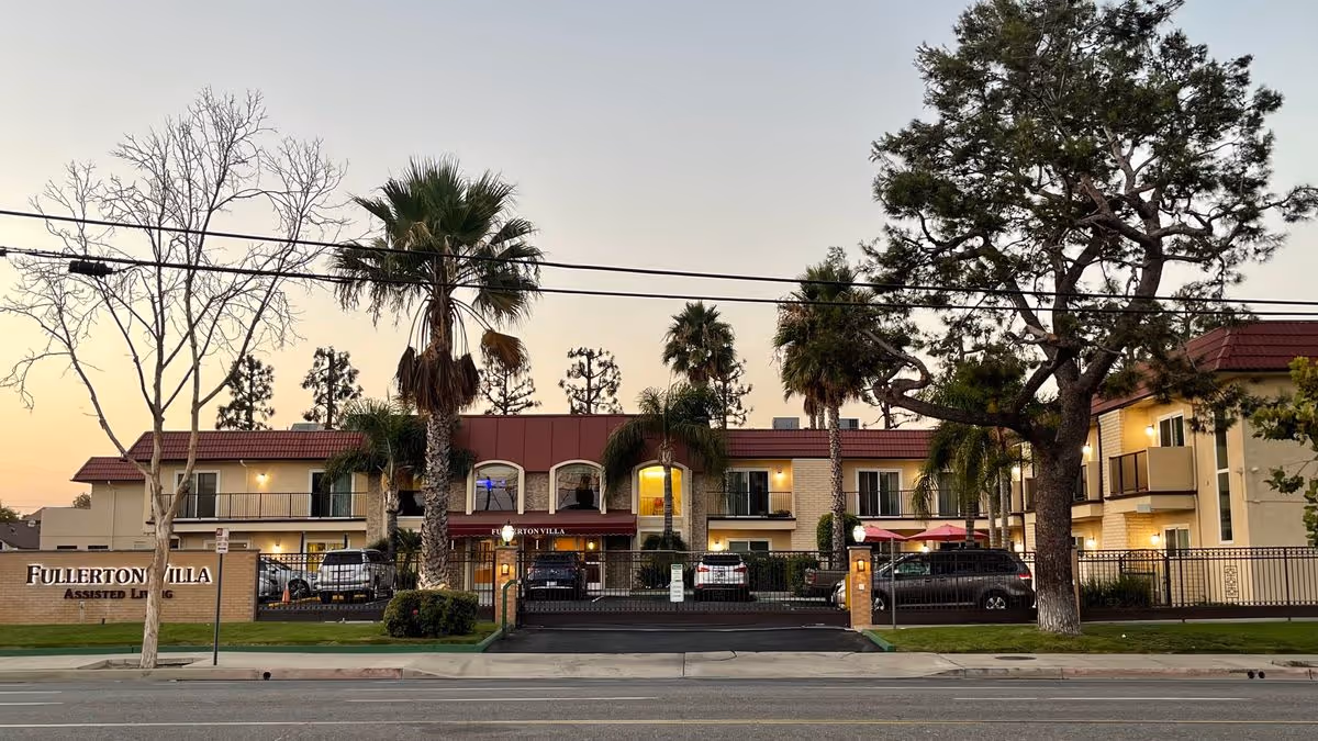 Two-story cream and red-roofed assisted living building labeled "Fullerton Villa" with a gated entrance, palm trees, and parked cars at dusk.