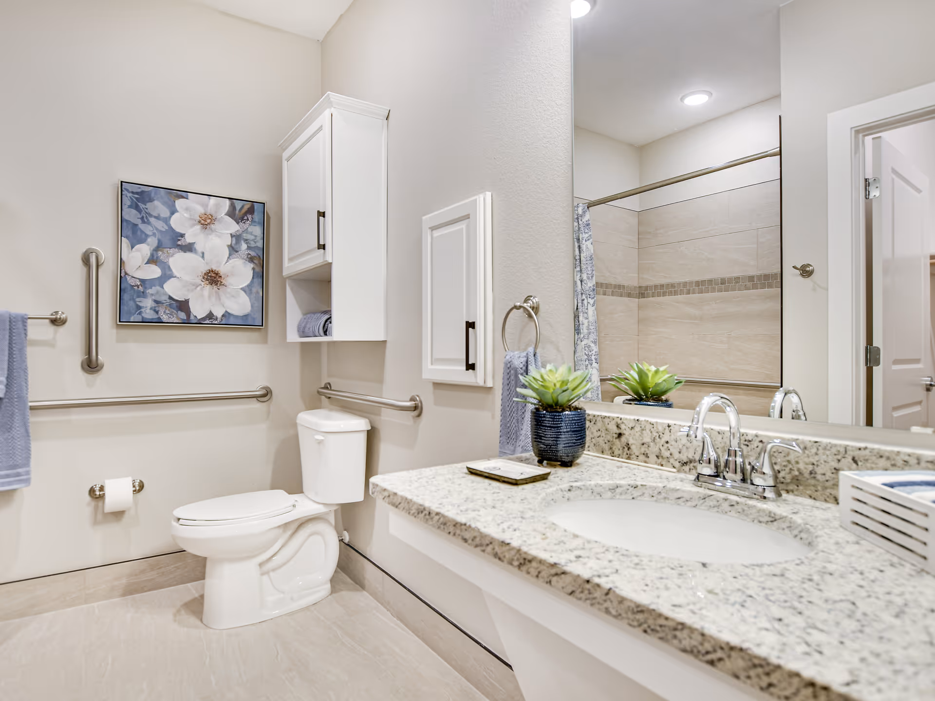 A clean and modern bathroom with a white toilet, granite countertop with an under-mount sink, and a large mirror above the sink. The bathroom features beige tiled floors and walls, a shower with a curtain, and stainless steel grab bars for accessibility. There is a white wall cabinet above the toilet, a towel ring with a blue towel, a small potted plant on the countertop, and a framed floral artwork on the wall.