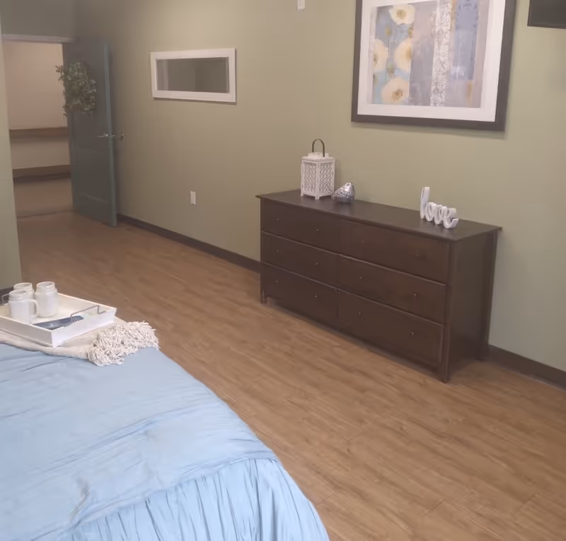 A bedroom with a light blue bedspread partially visible in the foreground. On the bed is a white tray holding two white mugs and a book. The room has wooden flooring and light green walls. Against the wall is a dark wooden dresser with six drawers, decorated with a white lantern, a silver bird figurine, and a white decorative word 'love'. Above the dresser hangs a framed floral artwork. An open door with a wreath on it leads to another room.
