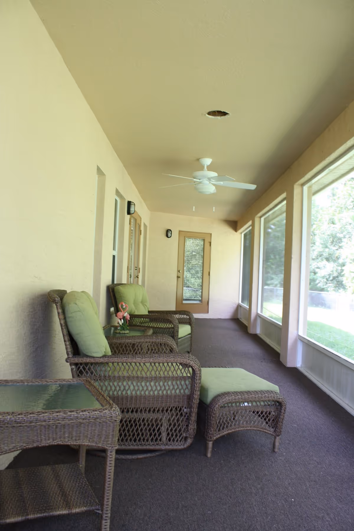 A screened-in porch area with wicker furniture including two armchairs with green cushions, a matching ottoman, and a glass-top side table. The porch has beige walls, a ceiling fan, and large screened windows overlooking a grassy outdoor area.