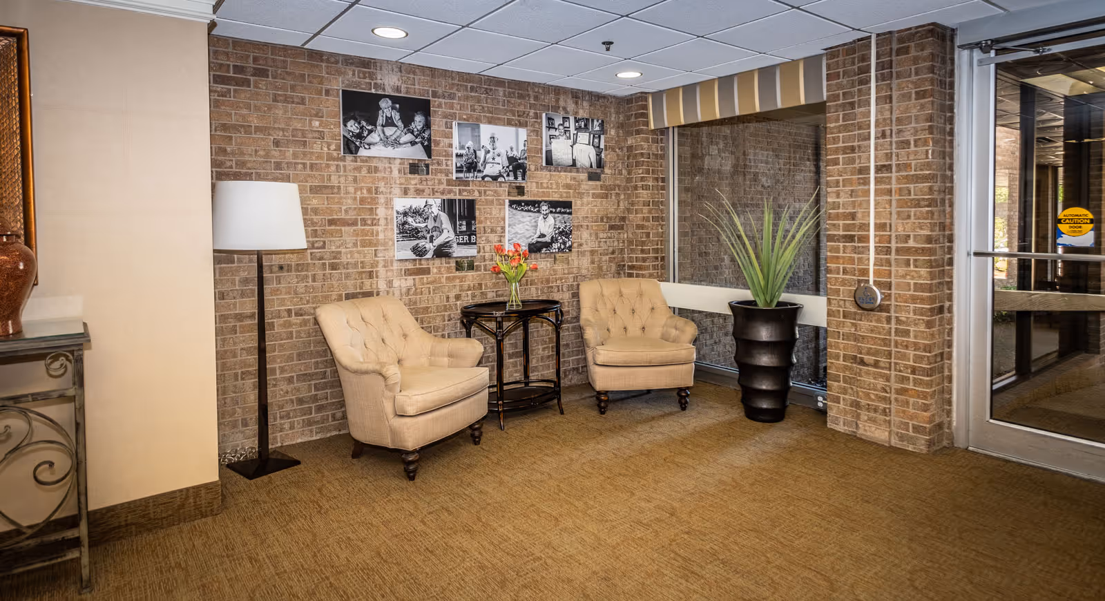 A cozy seating area in a nursing facility with two beige armchairs, a small dark wooden table with a vase of flowers, a tall floor lamp, and a large potted plant. The background features a brick wall adorned with black and white photographs. There is a glass door to the right leading outside.