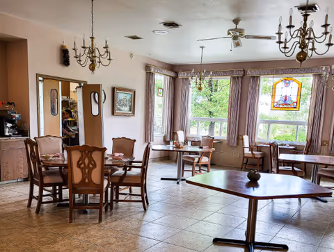 Bright dining room with wooden tables and chairs, chandeliers, large windows with purple curtains and a stained-glass panel.