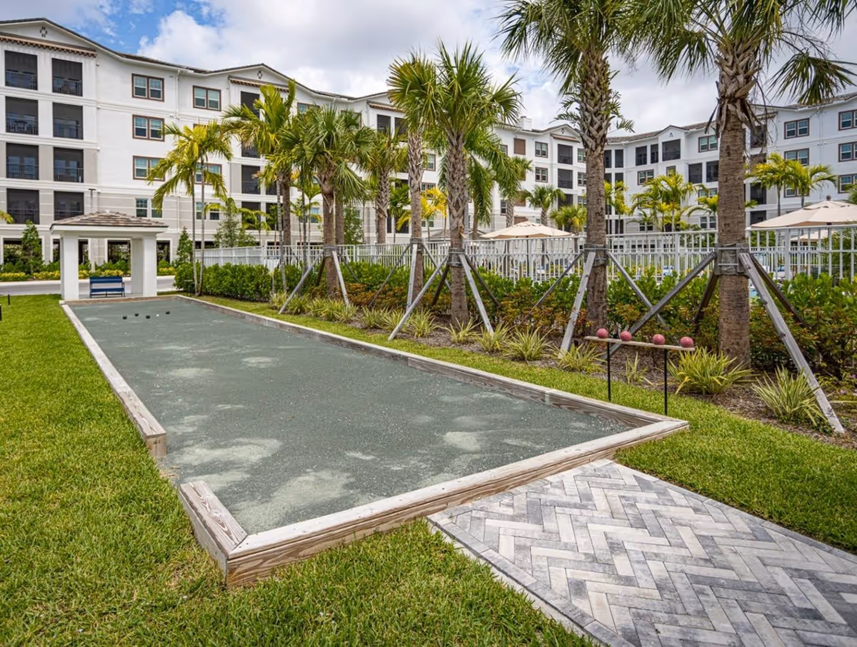 Outdoor bocce ball court with green playing surface surrounded by grass and palm trees, with a multi-story white residential building in the background under a partly cloudy sky.
