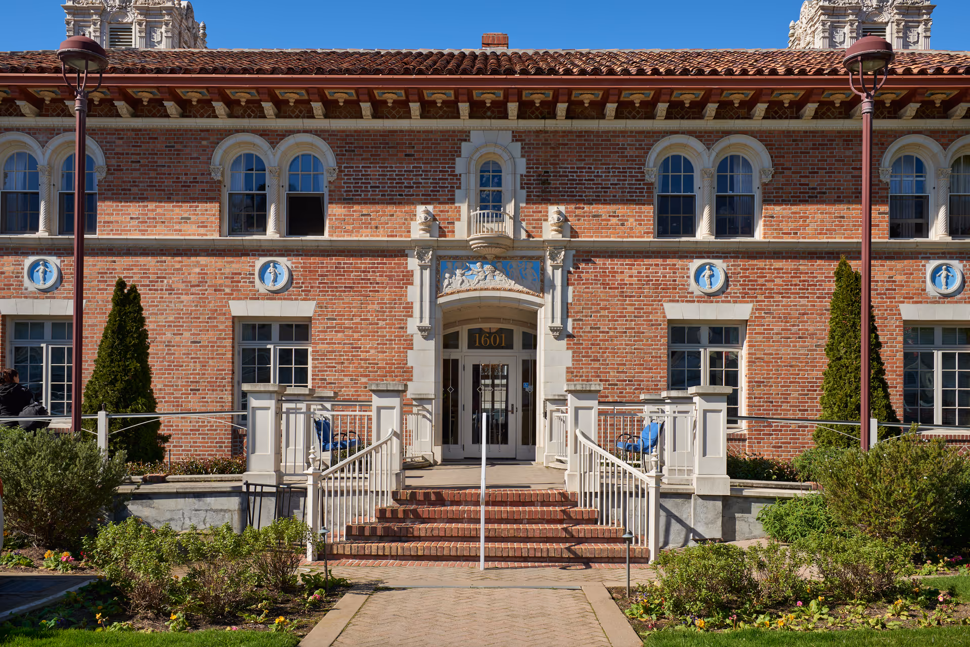 Front exterior view of a two-story brick building with arched windows and decorative stone accents. The entrance has a set of brick stairs with white railings leading to double glass doors under an ornate stone archway. There are two tall lampposts on either side of the walkway and well-maintained landscaping with bushes and flowers in front of the building.