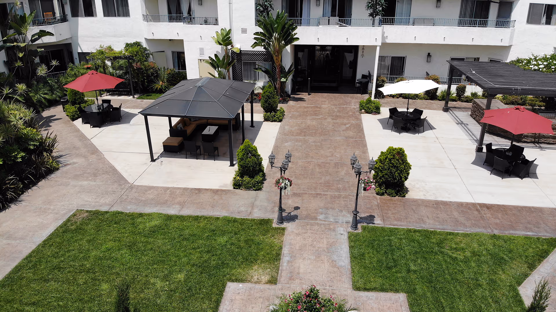 Outdoor courtyard area of Coronado Retirement Village featuring green lawns, paved walkways, several seating areas with tables and umbrellas, a covered gazebo with seating, and surrounding white multi-story building with balconies.
