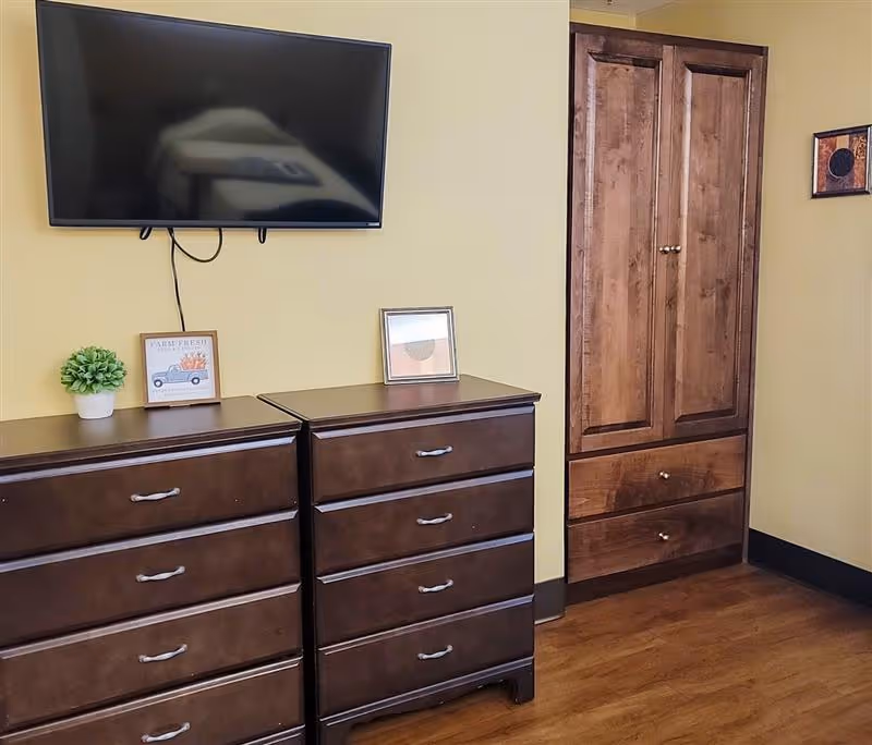 A room with two dark wooden dressers side by side, a mounted flat-screen TV above them, and a tall wooden cabinet with two drawers on the right. The walls are painted yellow, and there are small framed pictures and a potted plant on top of the dressers. The floor is wooden.