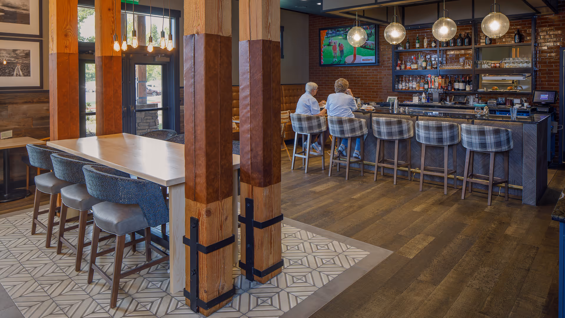 Interior view of a cozy bar and dining area with wooden floors and walls. There are two elderly women sitting on plaid bar stools at the bar counter, which is stocked with various bottles and glasses. A television is mounted on the brick wall behind the bar, showing a sports game. A high dining table with cushioned chairs is positioned near wooden pillars wrapped with leather accents. Pendant lights hang from the ceiling, illuminating the space.