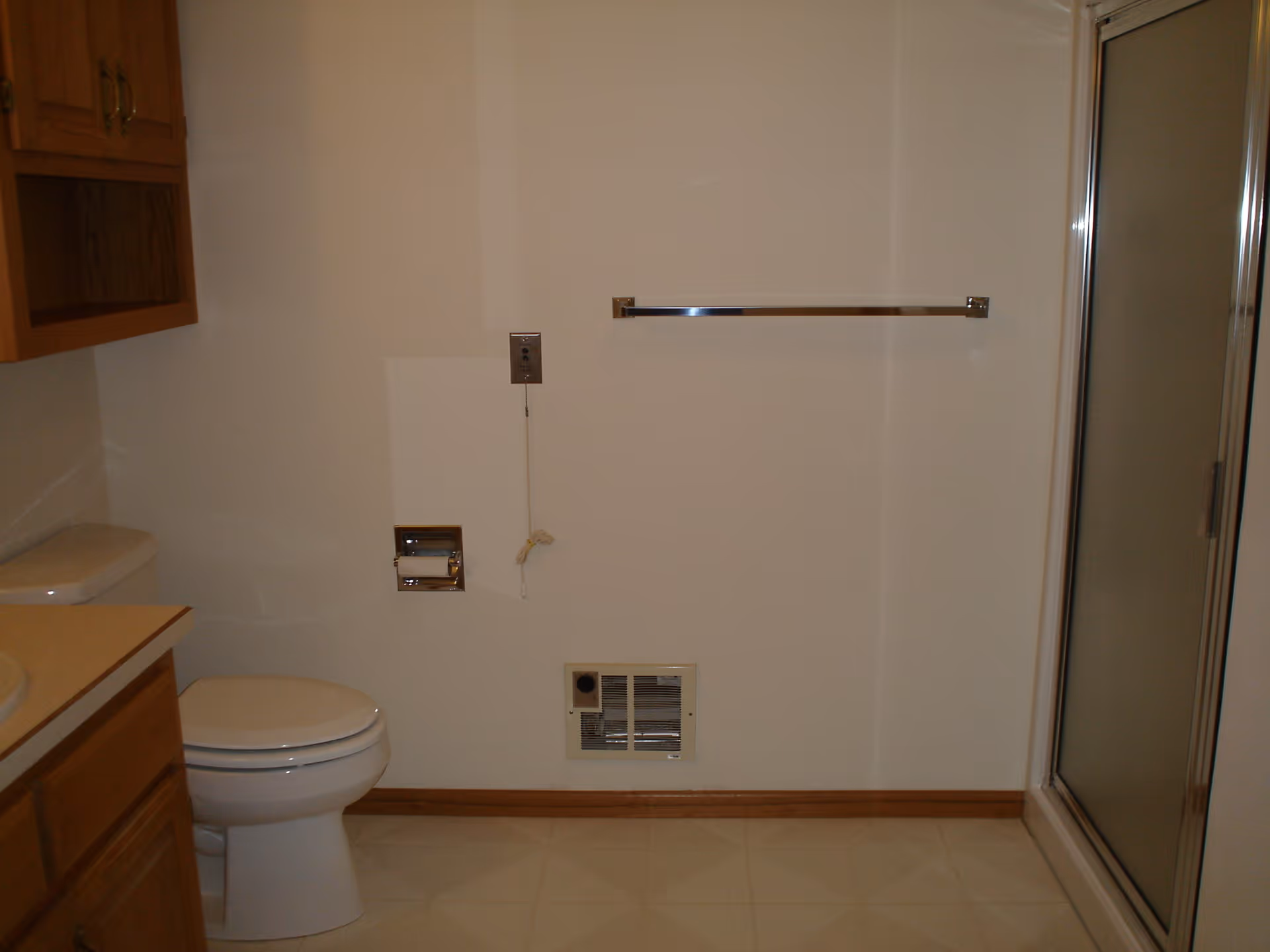 A bathroom with a white toilet, wooden cabinets, a towel rack on the wall, a toilet paper holder, a ventilation grate near the floor, and a glass shower door on the right side.