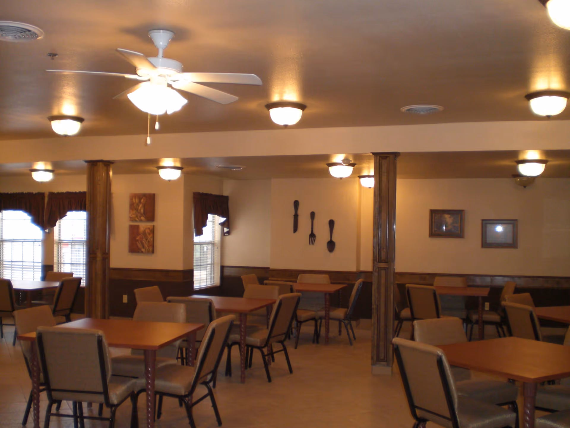 Interior view of a dining room with multiple tables and chairs arranged neatly. The room has beige walls, wooden wainscoting, and several ceiling lights along with a ceiling fan. There are windows with brown curtains and wall decorations including large fork, knife, and spoon sculptures and framed pictures.