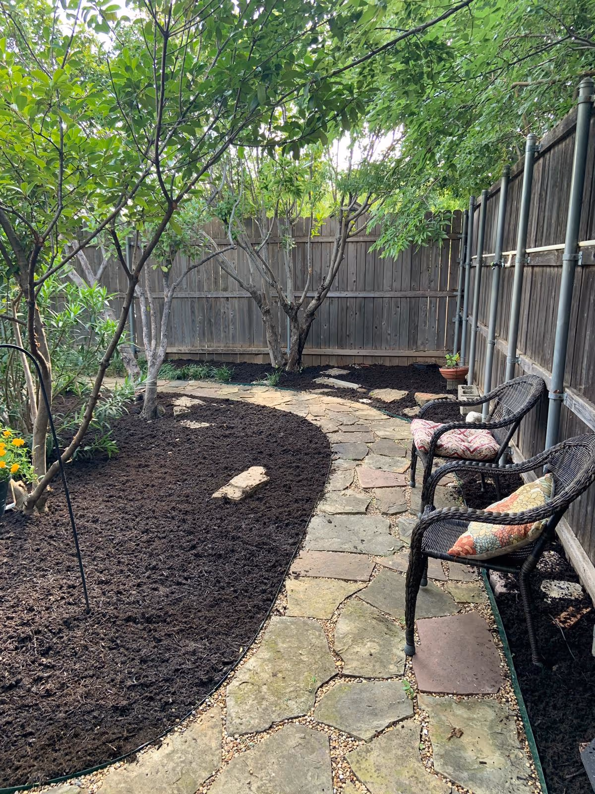 A small outdoor garden area with a curved stone pathway bordered by freshly mulched soil beds. There are several trees and plants along the left side, and two black wicker chairs with colorful cushions are placed on the right side next to a wooden fence.