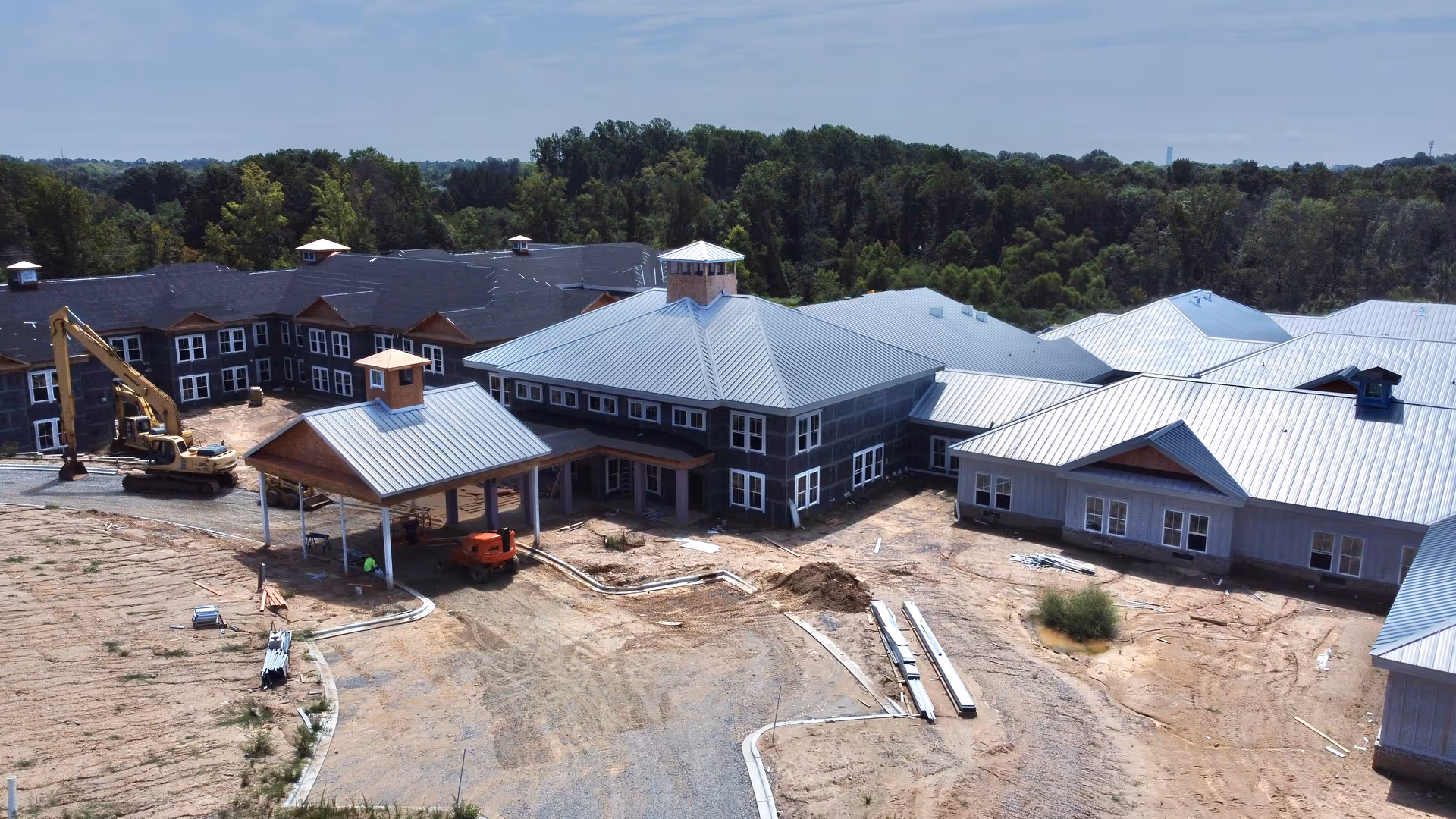 Aerial view of Arbor Ridge at Huntersville senior living facility under construction, showing multiple connected buildings with metal roofs, construction equipment, and cleared land surrounding the site with trees in the background.