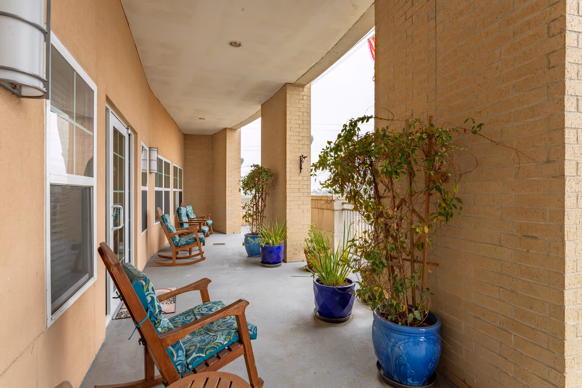 Covered outdoor patio area with several wooden rocking chairs featuring blue patterned cushions. The patio has beige brick walls and multiple potted plants in blue pots along the edge. The ceiling is white with recessed lighting.