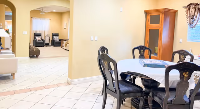 Interior view of a senior living facility showing a dining area with a round table covered with a white tablecloth and six dark wooden chairs. In the background, there is a living room area with two recliner chairs, a sofa, and a bed partially visible through an arched doorway. The floor is tiled, and there is a wooden cabinet against the wall near a window with curtains.