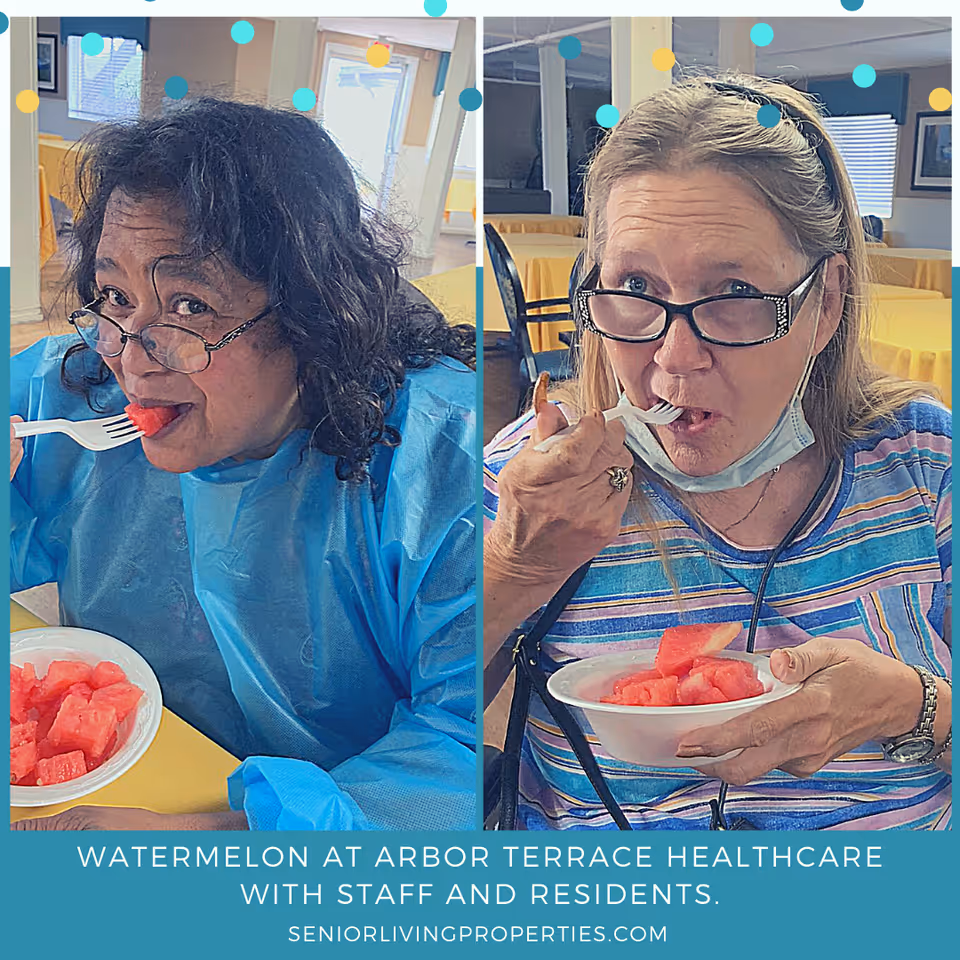 Two women eating watermelon from white bowls using plastic forks in a dining area with yellow tablecloths and chairs in the background. Both women are wearing glasses, one is in a blue protective gown and the other in a striped shirt with a face mask pulled down below her chin.